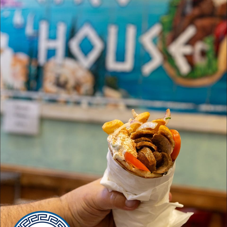 Hand holding a Gyro House gyro with meat, fries, sauce, and tomatoes, against a blue background.