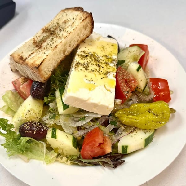 A Greek salad with feta cheese, olives, tomatoes, cucumbers, and grilled bread on a white plate.