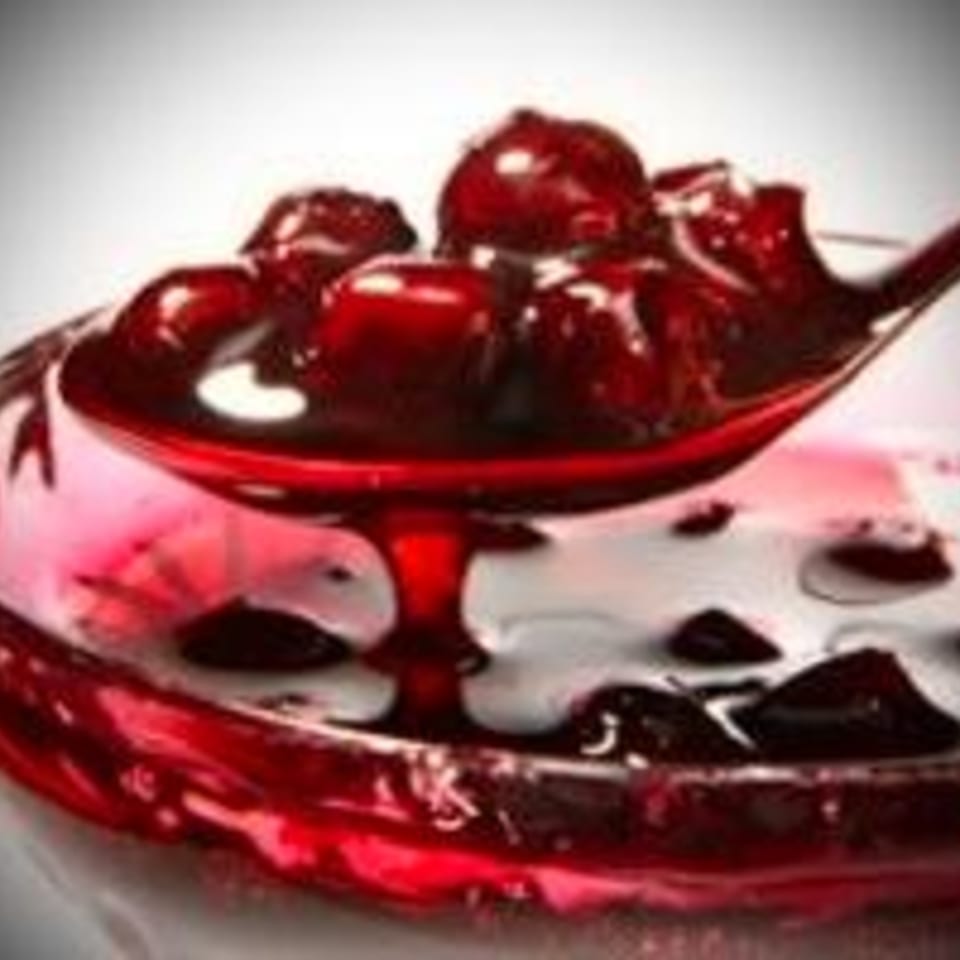 A spoon lifting dark red sour cherry preserves from a glass bowl, with syrup dripping.