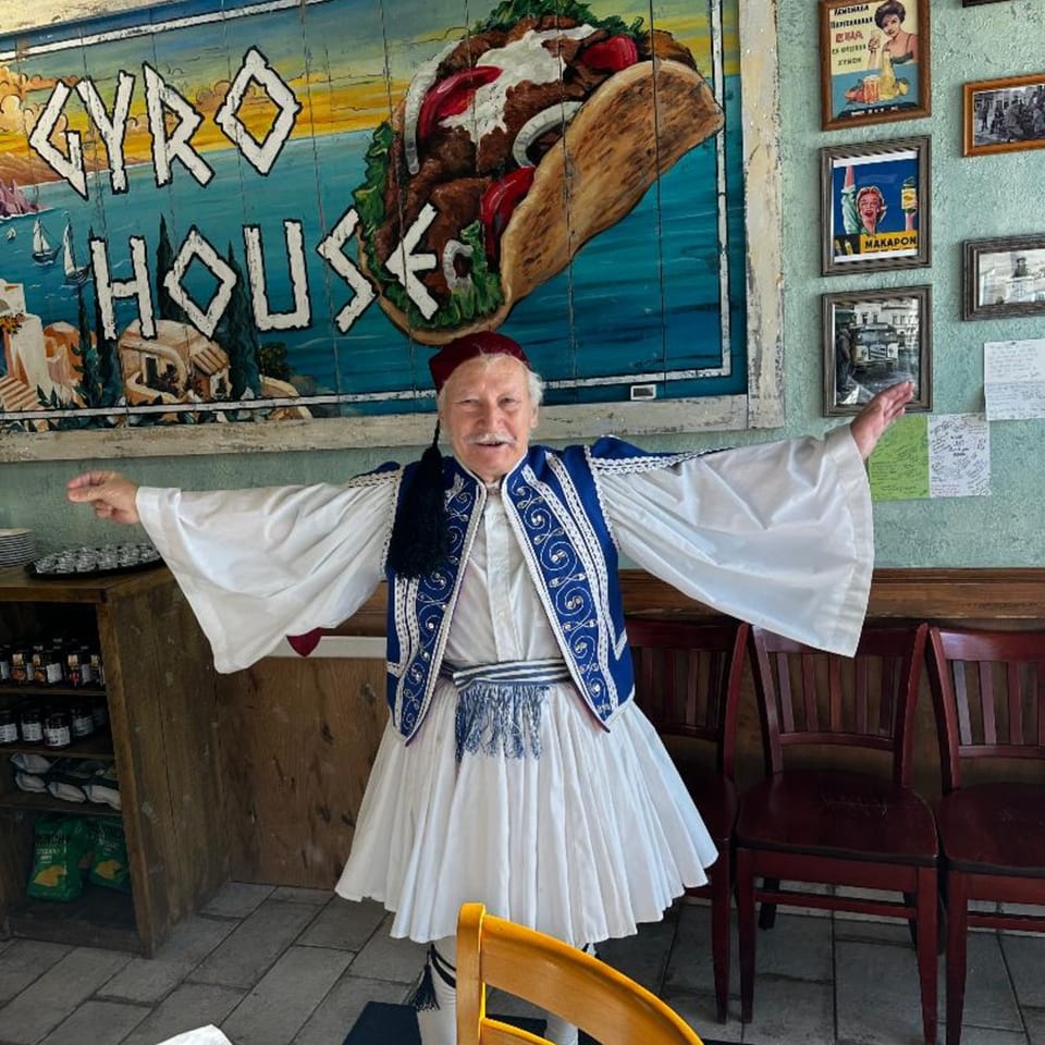 A man in traditional Greek attire stands before a large Gyro House mural of a gyro and a Greek village.
