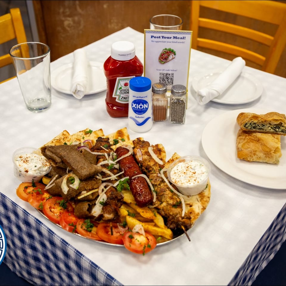 A large Greek platter with gyro meat, skewers, fries, tomatoes, pita, and tzatziki on a checkered tablecloth.