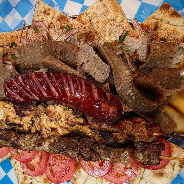 A large platter of Greek gyro meat, sausage, chicken, and beef skewers with pita bread and tomatoes.