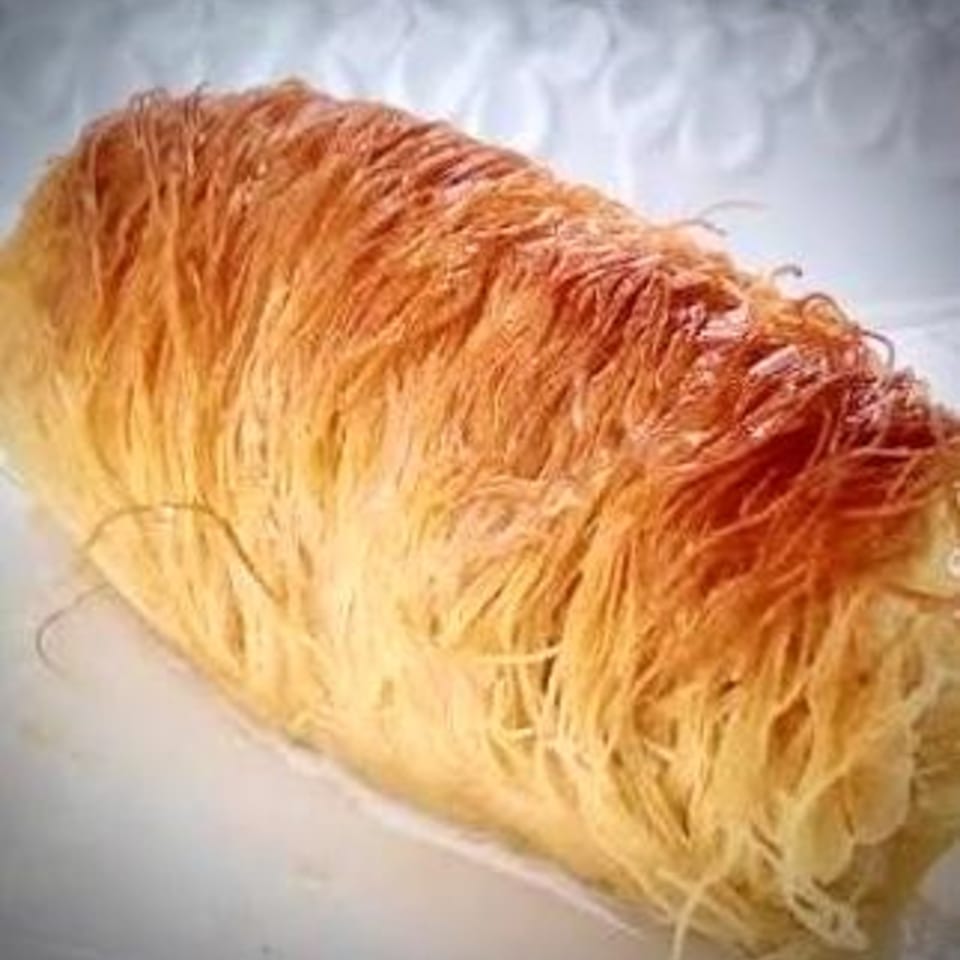 A golden-brown, cylindrical Greek dessert, possibly kataifi, with shredded pastry on a white surface.