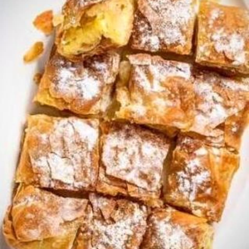 Close-up of golden brown Greek bougatsa pastries, dusted with powdered sugar, on a white plate.