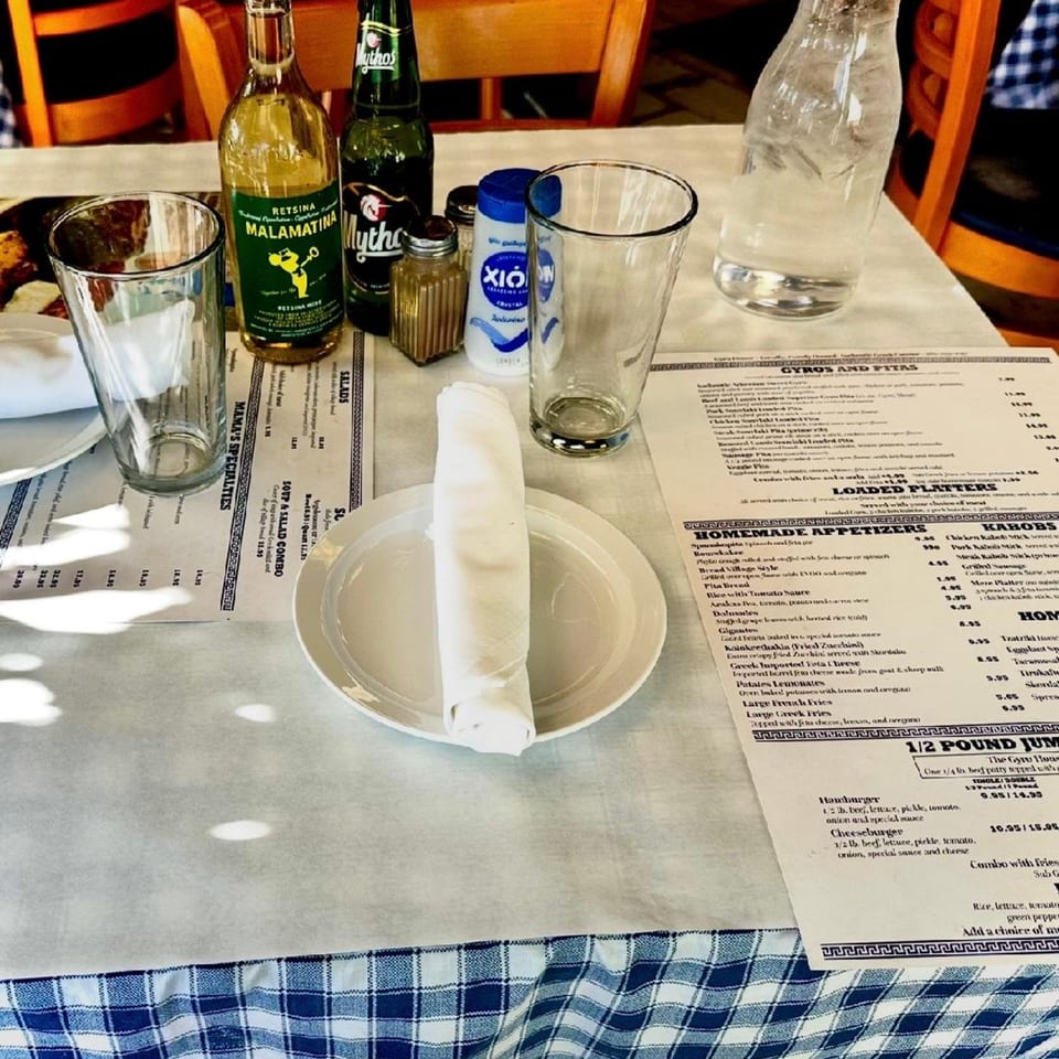 A Gyro House table set with Greek drinks, menus, and a white plate with a rolled napkin.